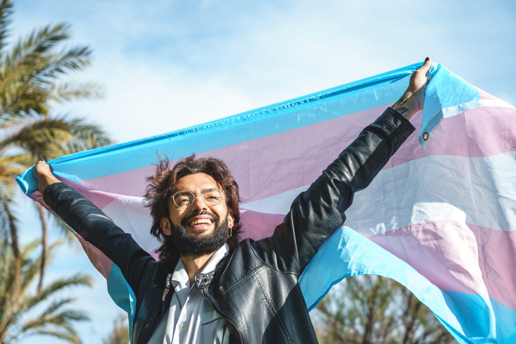 Low angle of cheerful young bearded man in casual clothes raising arms and smiling while standing on background of blue sky