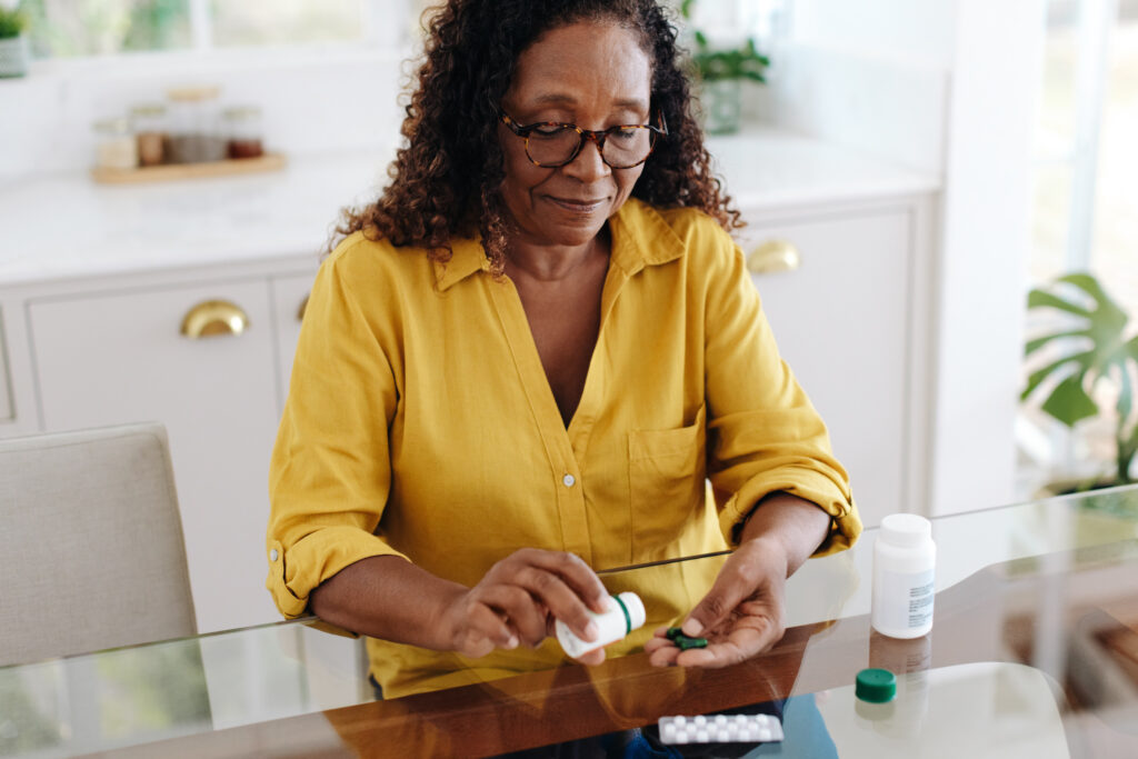 Black woman with a chronic health condition sits at her kitchen table taking her medication. Senior woman adhering to her prescribed treatment in order to efficiently manage her personal health.